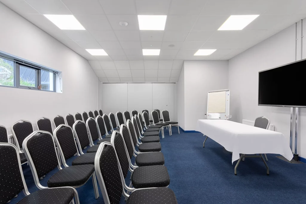 Meeting room with rows of black chairs, a white table, and presentation equipment, suitable for group discussions or events.